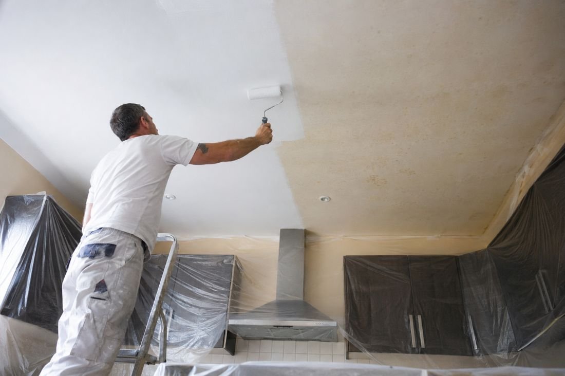 Pintor sobre escalera cubriendo con blanco el techo de una cocina manchado de grasa, con los muebles protegidos con plástico